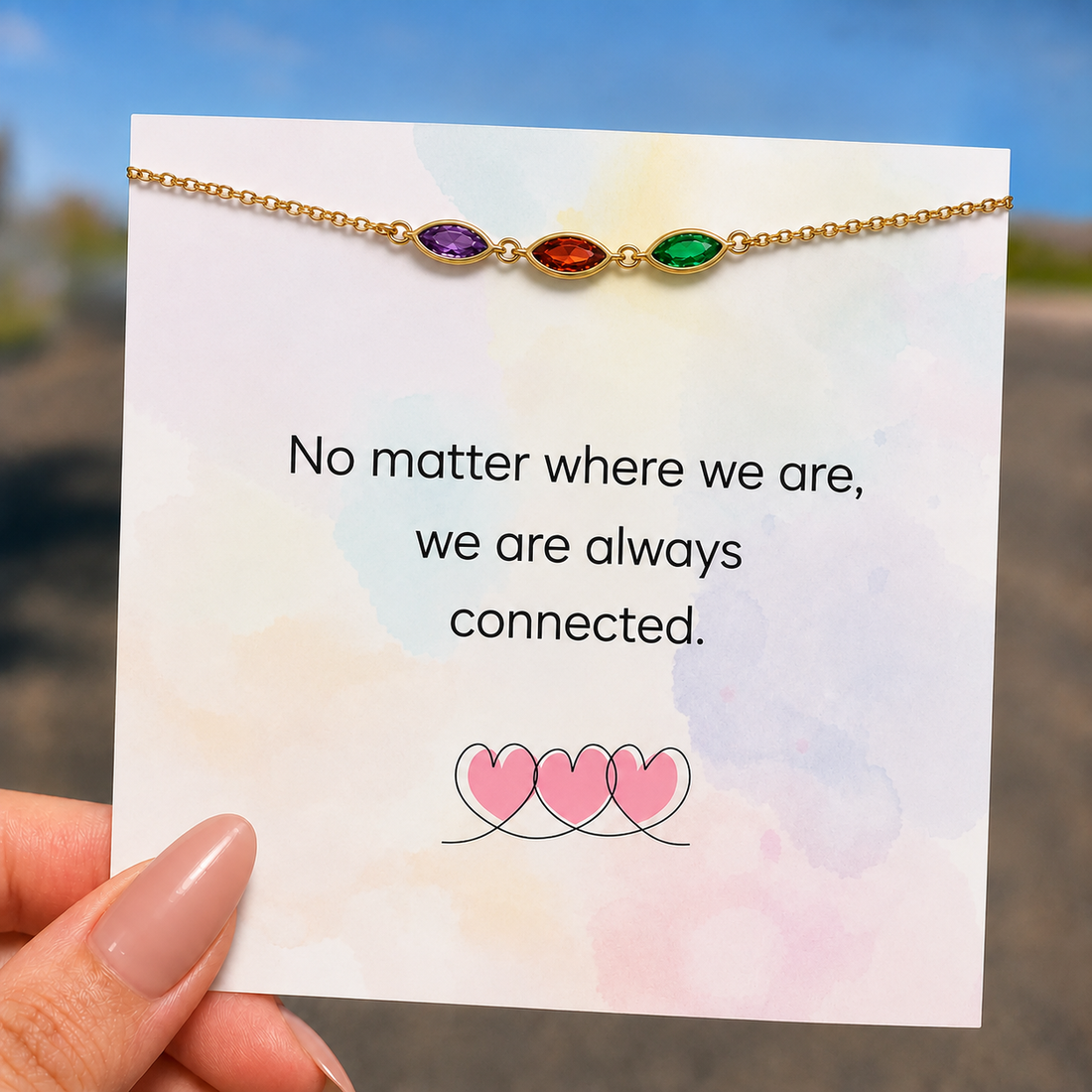 A gold chain bracelet with three small colored stones (purple, red, and green) displayed on a white card that reads 'No matter where we are, we are always connected.' A hand is holding the card, with a blurred outdoor background.