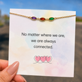 A gold chain bracelet with three small colored stones (purple, red, and green) displayed on a white card that reads 'No matter where we are, we are always connected.' A hand is holding the card, with a blurred outdoor background.